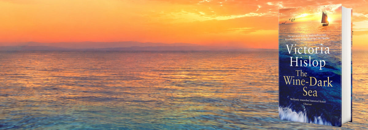Book cover for 'The Wine-Dark Sea' by Victoria Hislop, shown standing upright against a sunset ocean background. The cover features a blue sea with white surf and a small sailboat, with the title and author name in white text.